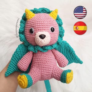 May include: A handmade pink crochet lion doll with a teal mane and cape, yellow horns, and black eyes. The doll has a beige muzzle and yellow and teal accents on its feet. The doll is sitting on a white surface.