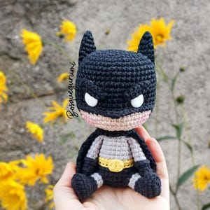 May include: A handmade crochet Batman doll. The doll is primarily black with a gray torso, a yellow belt, and white eyes. The doll has a small cape and is sitting. The background includes yellow flowers and a gray wall.