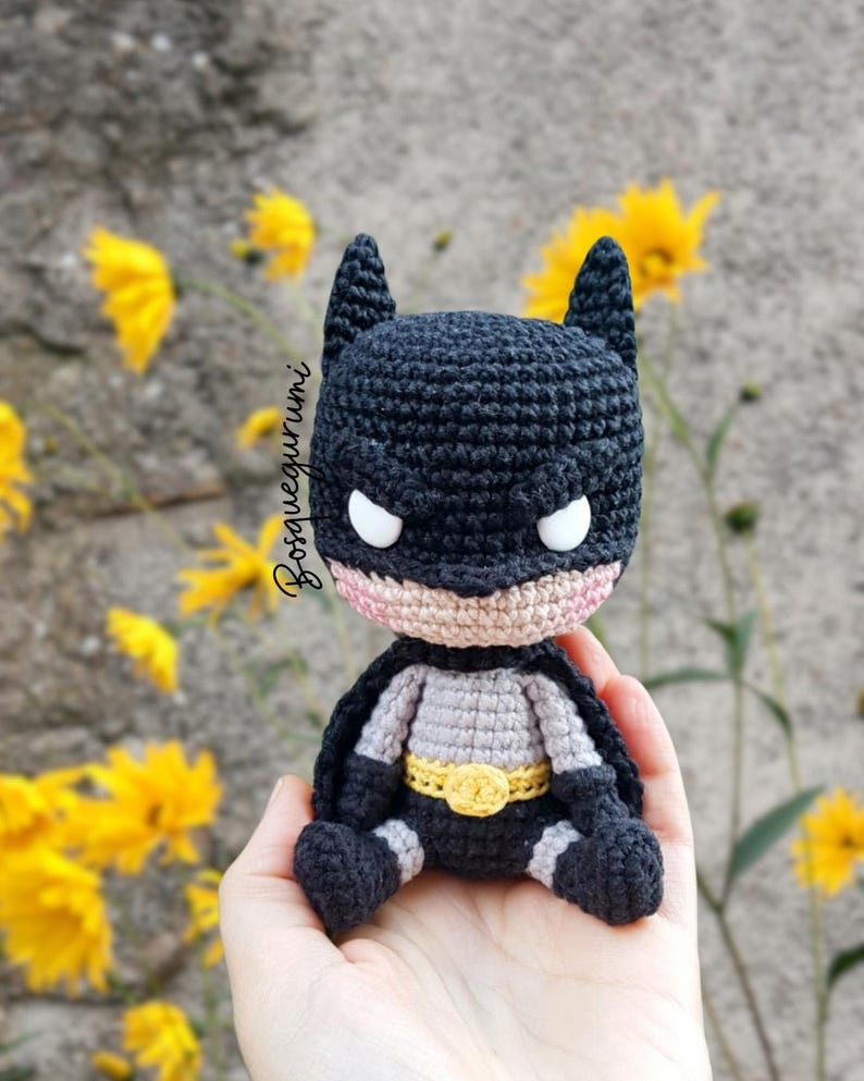 May include: A handmade crochet Batman doll. The doll is primarily black with a gray torso, a yellow belt, and white eyes. The doll has a small cape and is sitting. The background includes yellow flowers and a gray wall.