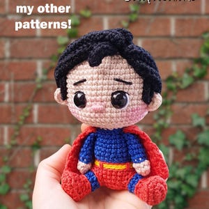 May include: A handmade crochet Superman doll with a red cape, blue and red suit, and black hair. The doll is sitting and has large black eyes. Text on the image reads "Check out my other patterns!" and "@BOSQUEGURUMI".
