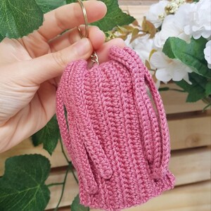 May include: A handmade, pink crochet keychain pouch with a gold-colored ring. The pouch has a ribbed texture and two straps. The text "@BOSQUEGURUMI" is visible in the bottom right corner. The background includes greenery and white flowers.