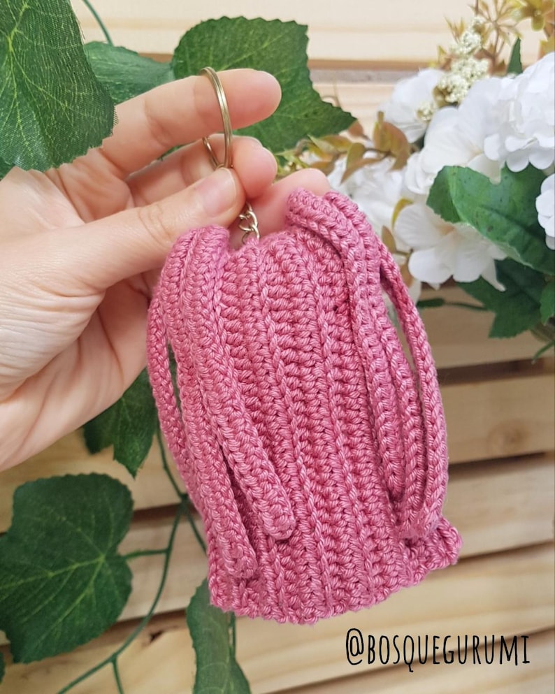 May include: A handmade, pink crochet keychain pouch with a gold-colored ring. The pouch has a ribbed texture and two straps. The text "@BOSQUEGURUMI" is visible in the bottom right corner. The background includes greenery and white flowers.