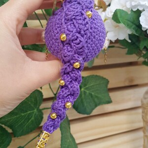 May include: A handmade, purple crochet ornament with gold accents. The ornament has a bulbous top with a braided design and a long, twisted tail. Gold beads are evenly spaced along the tail. The ornament is held by a gold ring.