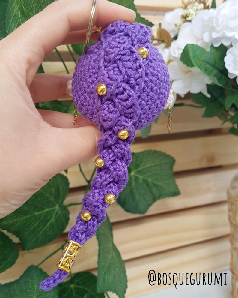 May include: A handmade, purple crochet ornament with gold accents. The ornament has a bulbous top with a braided design and a long, twisted tail. Gold beads are evenly spaced along the tail. The ornament is held by a gold ring.