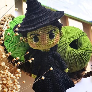 以下が含まれることがあります： 黒い帽子とドレス、緑色の肌、大きな黒い目をした手作りの編み物魔女人形。 人形は、緑色の糸とドライフラワーが入った織りバスケットに飾られています。 Instagramハンドル@BOSQUEGURUMIが表示されます。