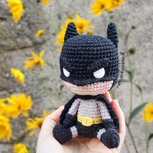 May include: A handmade crochet Batman doll, featuring a black cowl with pointed ears, white eyes, and a pink mouth. The doll has a gray and black suit with a yellow belt. The background includes yellow flowers and a gray wall.