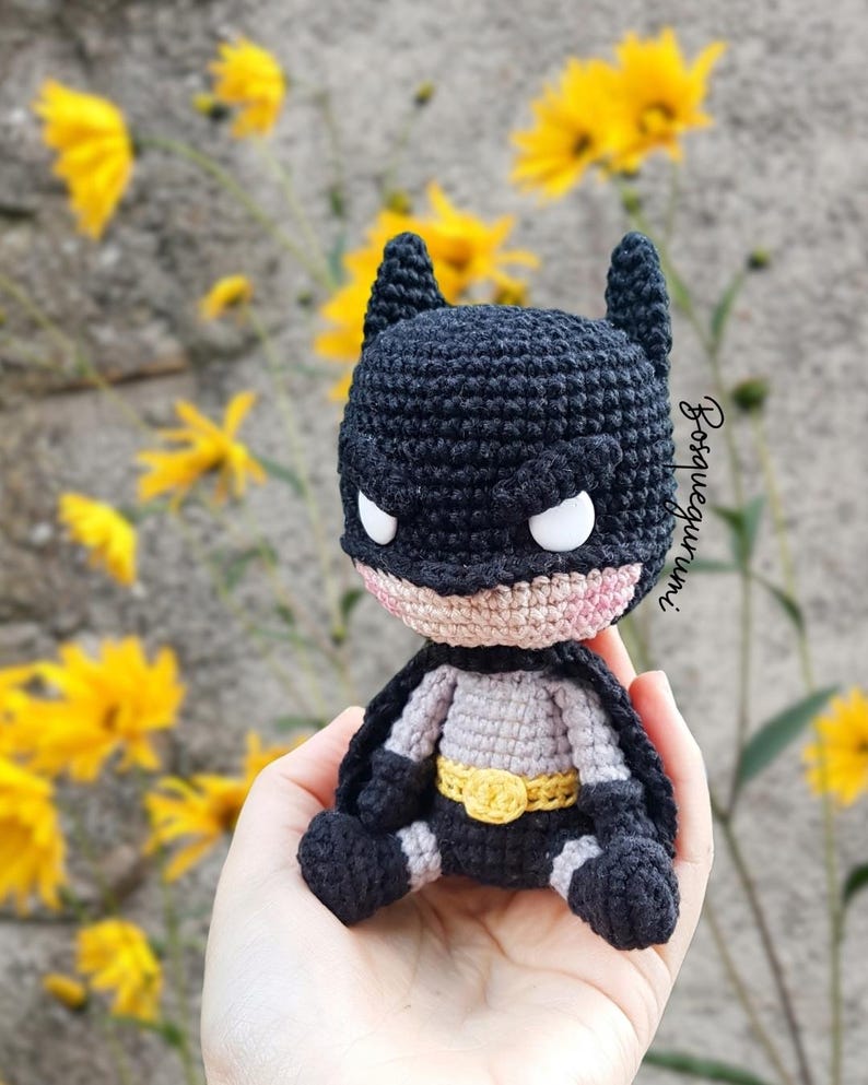 May include: A handmade crochet Batman doll, featuring a black cowl with pointed ears, white eyes, and a pink mouth. The doll has a gray and black suit with a yellow belt. The background includes yellow flowers and a gray wall.