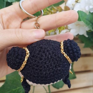 May include: A handmade black crochet keychain featuring a doll with two buns, gold accents, and a white face. The keychain is attached to a gold-colored ring. The text "@BOSQUEGURUMI" is visible in the bottom right corner.