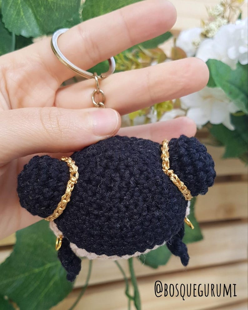 May include: A handmade black crochet keychain featuring a doll with two buns, gold accents, and a white face. The keychain is attached to a gold-colored ring. The text "@BOSQUEGURUMI" is visible in the bottom right corner.