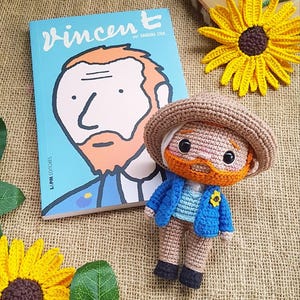 May include: A crocheted amigurumi doll of Vincent van Gogh, wearing a blue jacket, brown hat, and orange beard. The doll is sitting on a burlap surface next to a book titled "Vincent" by Barbara Stok. There is a yellow crocheted sunflower in the background.
