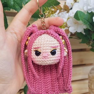 May include: A handmade pink crochet doll keychain with a gold-colored keyring. The doll has a light skin tone, black eyes, and pink hair. The keychain is a unique, handcrafted accessory.