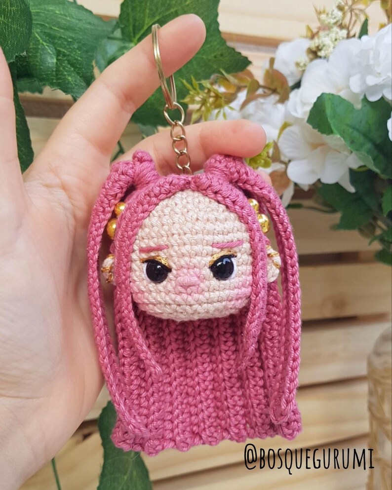 May include: A handmade pink crochet doll keychain with a gold-colored keyring. The doll has a light skin tone, black eyes, and pink hair. The keychain is a unique, handcrafted accessory.