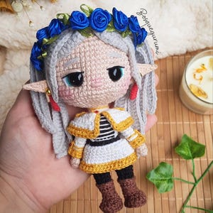 Könnte beinhalten: Handgemachte Häkelpuppe mit grauem Haar, blauem Blumenkranz und Elfenohren. Die Puppe trägt ein weißes Kleid mit goldenen Verzierungen und ein schwarz-weiß gestreiftes Oberteil. Braune Stiefel vervollständigen das Outfit.