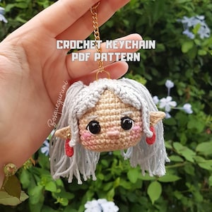 Puede incluir: Un llavero de ganchillo con la cara de una muñeca con pelo gris, mejillas rosadas y pendientes rojos. El llavero tiene una cadena dorada y el texto "CROCHET KEYCHAIN PDF PATTERN" está visible.