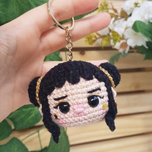 May include: Crocheted keychain featuring a doll face with black hair styled in two buns, gold accents, and a star-shaped embellishment. The doll has large black eyes and rosy cheeks. The keychain is held by a gold ring and chain. The text "@BOSQUEGURUMI" is visible.