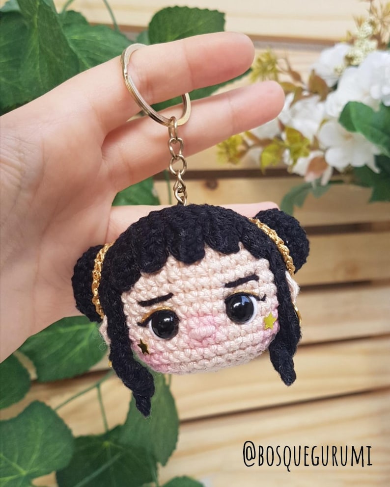 May include: Crocheted keychain featuring a doll face with black hair styled in two buns, gold accents, and a star-shaped embellishment. The doll has large black eyes and rosy cheeks. The keychain is held by a gold ring and chain. The text "@BOSQUEGURUMI" is visible.