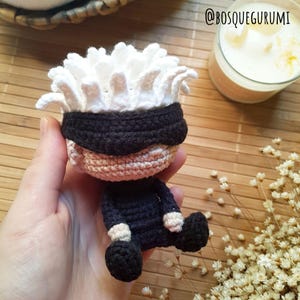 May include: A handmade crochet doll featuring a character with white hair, a black blindfold, and a dark blue outfit. The doll is seated with its legs bent. The text "@BOSQUEGURUMI" is visible in the upper right corner.