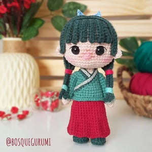 Puede incluir: Una muñeca de crochet que lleva un vestido verde y blanco con una falda roja. La muñeca tiene el pelo largo y negro con cintas azules y grandes ojos marrones.