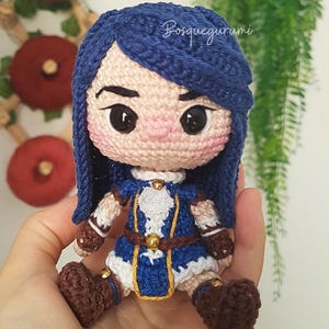 Pode incluir: Boneca de croch&ecirc; com cabelo azul vestindo um traje azul e branco com detalhes dourados. A boneca tem sapatos marrons e est&aacute; segurando uma pequena espada.