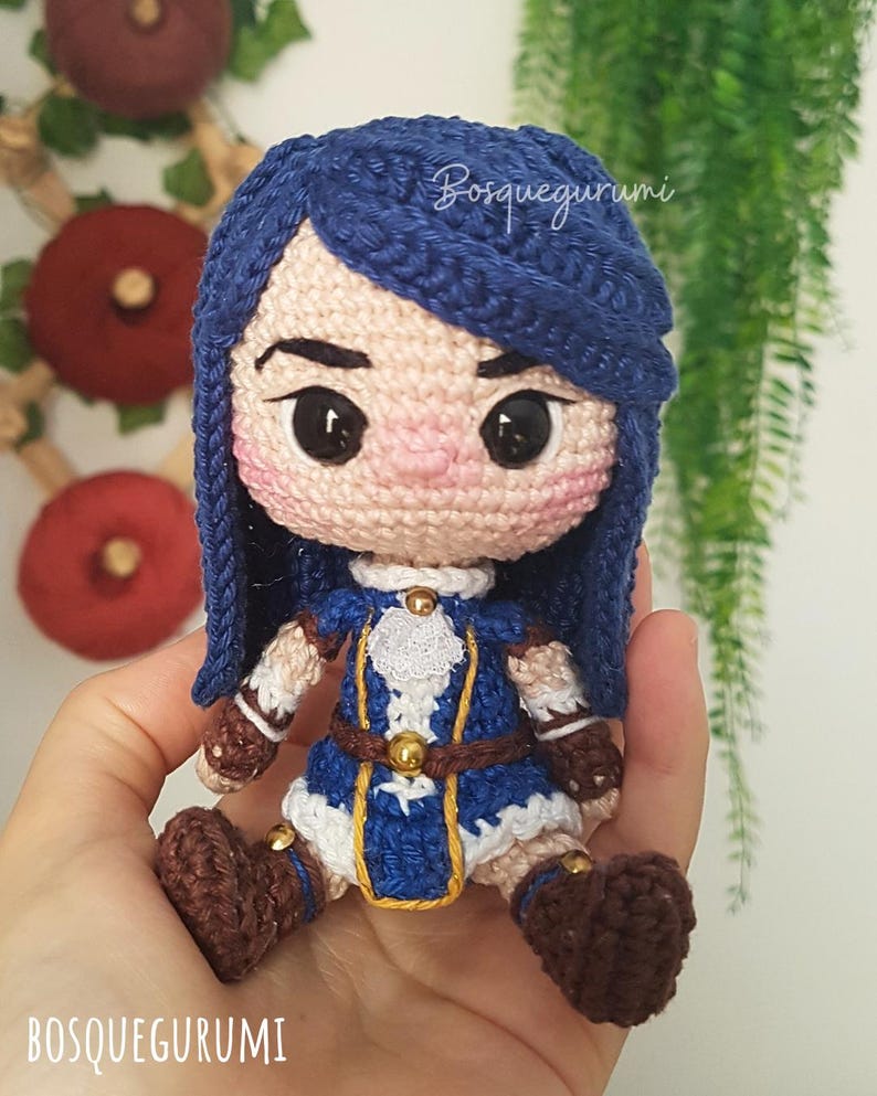 Pode incluir: Boneca de croch&ecirc; com cabelo azul vestindo um traje azul e branco com detalhes dourados. A boneca tem sapatos marrons e est&aacute; segurando uma pequena espada.