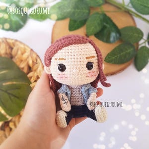 May include: A crocheted doll with brown hair, wearing a blue denim jacket, a pink shirt, and blue jeans. The doll has a small tattoo on its arm.