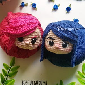 Pode incluir: Duas bonecas de croch&ecirc; com cabelo de l&atilde;, uma com l&atilde; vermelha e a outra com l&atilde; azul. As bonecas t&ecirc;m olhos grandes e est&atilde;o sentadas em uma superf&iacute;cie branca. O texto "BOSQUEGURUMI" &eacute; vis&iacute;vel na imagem.