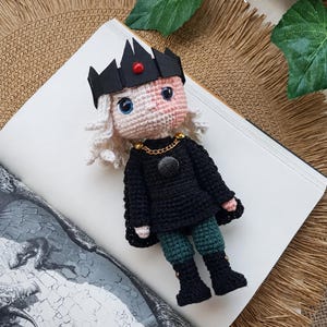 May include: A handmade crochet doll featuring white hair, a black crown with a red centre gem, and a black tunic with a gold chain detail. The doll wears dark green trousers and black boots, with a black cape attached to the shoulders.