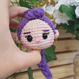 May include: Crocheted doll keychain featuring a woman's face with purple hair and gold earrings. The doll has black eyes, a pink nose, and a purple braid. The keychain is held by a gold chain.