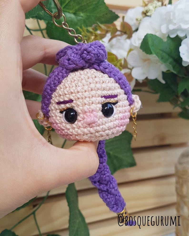 May include: Crocheted doll keychain featuring a woman's face with purple hair and gold earrings. The doll has black eyes, a pink nose, and a purple braid. The keychain is held by a gold chain.
