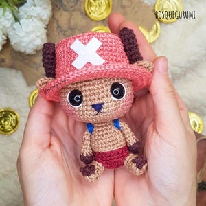 Może przedstawiać: Szydełkowa pluszowa zabawka przedstawiająca postać z kreskówki, trzymana w dłoniach. Zabawka ma różowy kapelusz z białym krzyżem, duże czarne oczy i mały niebieski nos. Postać ma na sobie czerwone szorty i niebieskie szelki. Widoczna jest nazwa marki "BOSQUEGURUMI".