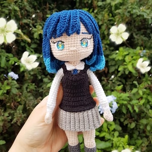 以下が含まれることがあります： 青い髪と青と金の目を持つ手作りのかぎ針編みの人形。人形は黒いベスト、白い襟付きシャツ、灰色のプリーツスカートを着ています。人形は、緑の葉と白い花の背景の前に保持されています。