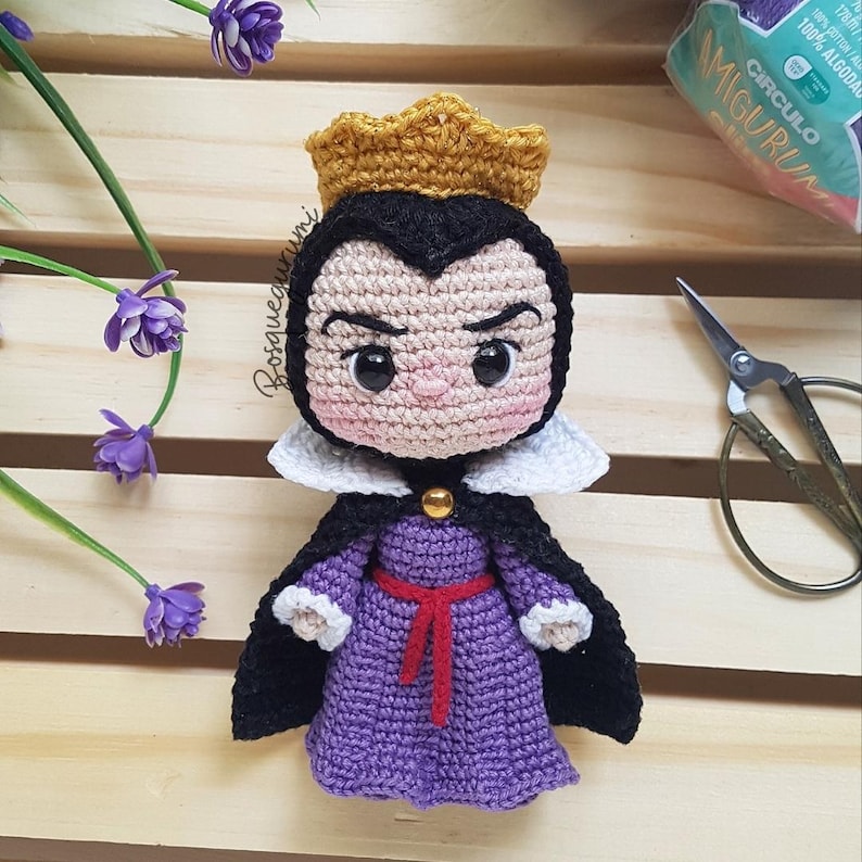 May include: Crocheted doll of a character wearing a gold crown, black cape, and purple dress with a red belt. The doll has a white collar and a stern expression. The doll is handmade and has the word "Bosquegurumi" on it.