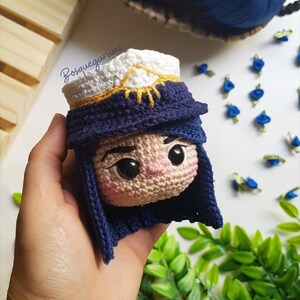 Pode incluir: Boneca amigurumi de croch&ecirc; com cabelo azul, um chap&eacute;u branco e dourado e um rosto detalhado. A boneca est&aacute; sendo segurada em uma m&atilde;o.