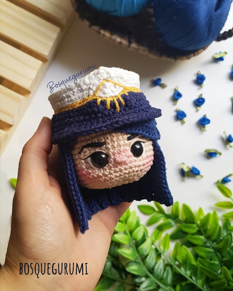 Pode incluir: Boneca amigurumi de croch&ecirc; com cabelo azul, um chap&eacute;u branco e dourado e um rosto detalhado. A boneca est&aacute; sendo segurada em uma m&atilde;o.