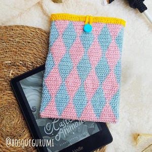 May include: A handmade crochet Kindle cover with a pink and light blue diamond pattern. The cover has a yellow border and a turquoise button closure. The Kindle is resting on a woven mat, with the text "Castle Animad" visible on the screen.