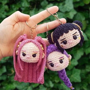 May include: Three handmade crochet doll keychains. One has pink hair, one has black hair, and one has purple hair. Each doll has a gold keychain ring and a detailed face. The background is a blurred green.