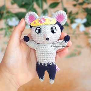 May include: A handmade crochet mouse doll with a yellow headband and a pink gem. The mouse has large black eyes, gray and pink ears, and is wearing a dark blue outfit. The doll is holding its arms out and has the text "@bosquegurumi".