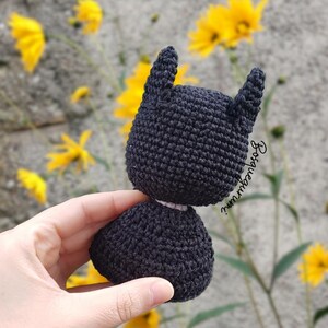 May include: A handmade black crochet Batman doll with pointed ears. The doll is held in front of a background of yellow flowers and a gray wall. The doll's head and body are separate pieces.