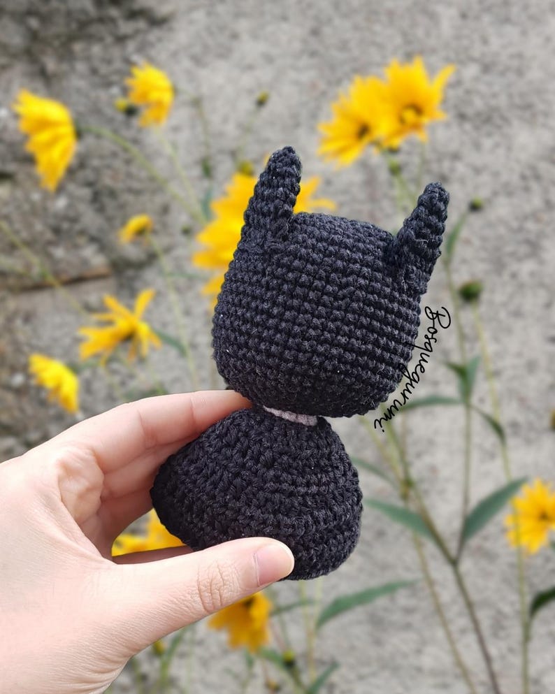 May include: A handmade black crochet Batman doll with pointed ears. The doll is held in front of a background of yellow flowers and a gray wall. The doll's head and body are separate pieces.