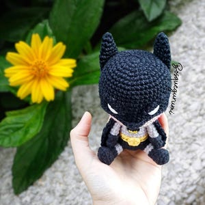 May include: A handmade, crocheted Batman doll in black, gray, and yellow. The doll features the iconic bat ears, a yellow belt, and a gray cape. The doll is held in a hand, with a yellow flower and green leaves in the background.
