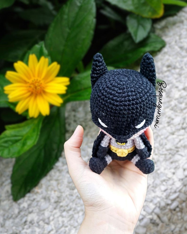 May include: A handmade, crocheted Batman doll in black, gray, and yellow. The doll features the iconic bat ears, a yellow belt, and a gray cape. The doll is held in a hand, with a yellow flower and green leaves in the background.
