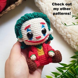 May include: A handmade crochet doll of the Joker character, featuring a white face with red and blue makeup, green hair, and a red suit. The doll is held in a hand. Text on the image reads "@BOSQUEGURUMI" and "Check out my other patterns!"