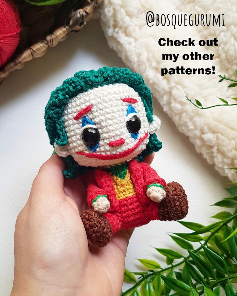 May include: A handmade crochet doll of the Joker character, featuring a white face with red and blue makeup, green hair, and a red suit. The doll is held in a hand. Text on the image reads "@BOSQUEGURUMI" and "Check out my other patterns!"