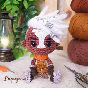 May include: A handmade crochet doll with a brown complexion, white hair, and an orange scarf. The doll is wearing a brown vest and grey trousers. The doll is sitting on a white surface. The text "Bosquegurumi" is written on the image.