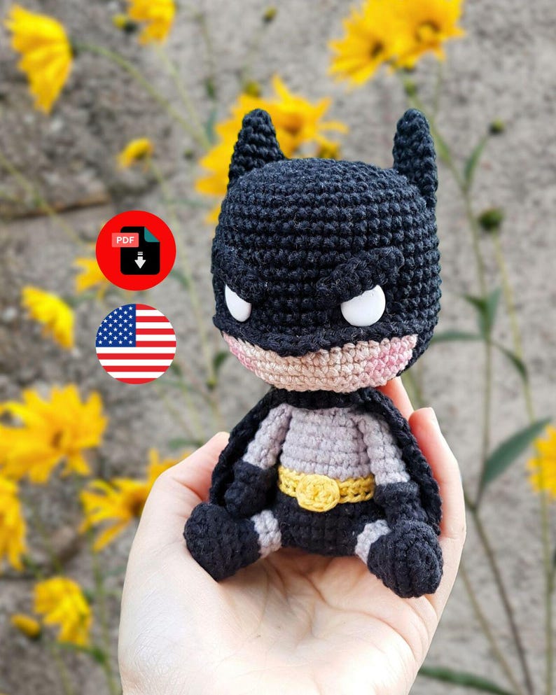 May include: A handmade crochet Batman doll, featuring a black cowl with white eye accents, a gray and black suit, and a yellow belt. The doll is held in a hand, with yellow flowers and a gray wall in the background. A PDF download icon and an American flag icon are also visible.