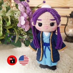 Puede incluir: Muñeca de ganchillo hecha a mano con cabello morado y un atuendo azul y marino. La muñeca tiene un sombrero blanco y está colocada frente a un arreglo floral. El atuendo de la muñeca tiene ribetes dorados.