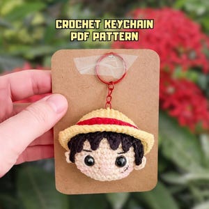 Può includere: Portachiavi all'uncinetto raffigurante il volto di un personaggio con cappello di paglia e capelli neri. Il portachiavi è attaccato a un anello rosso e a un supporto di cartone. Il testo "CROCHET KEYCHAIN PDF PATTERN" è visualizzato in alto.