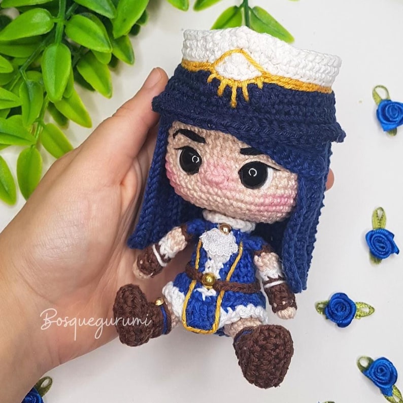 Pode incluir: Uma boneca de croch&ecirc; com cabelo azul, um traje azul e branco e sapatos marrons. A boneca est&aacute; usando um chap&eacute;u branco com um desenho de sol dourado.