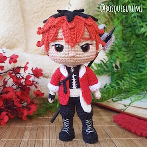 Op de afbeelding: Een handgemaakte gehaakte pop met rood haar en een rode jas met witte rand. De pop draagt een zwarte broek en laarzen met witte veters. De pop heeft een zwart zwaard en een klein paars en wit accessoire. De tekst "@BOSQUEGURUMI" staat rechtsboven.