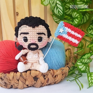 May include: A handmade crochet doll of a man with black hair, a mustache, and a football. He is holding a small Puerto Rican flag. The doll is in a woven basket with red and blue yarn. The doll is wearing a white outfit.